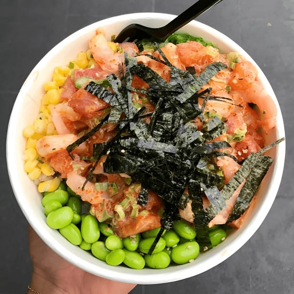 A Poke Chop poke bowl with salmon, shrimp, edamame, corn, and seaweed, held by a hand.
