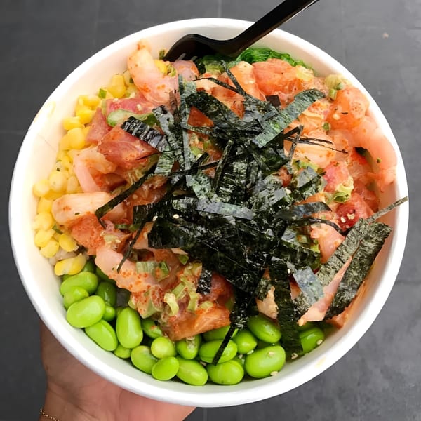 A Poke Chop poke bowl with salmon, shrimp, corn, edamame, and seaweed, held by a hand.